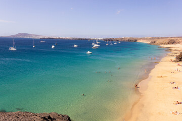Few people on Papagayo beach with sailboats over turquoise water sea in Lanzarote. Tourist bay on volcanic coast in Canary Islands on sunny day. Summer holidays, travel destination concepts