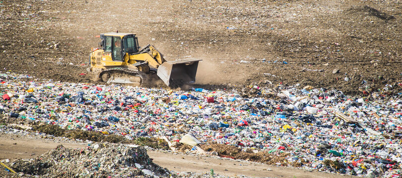 Yellow tractor works on huge landfill of big city. A lot of garbage on dump. Waste sorting on dumping site.