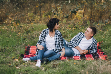 A loving guy and a girl in plaid shirts and jeans on a red plaid in the fall sit and hold hands on a background of green grass.