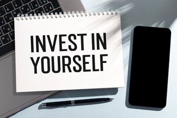 Text INVEST IN YOURSELF on card with laptop and phone on office desk. Flat lay, top view.