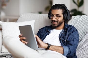 Handsome Indian Guy In Wireless Headphones Relaxing With Digital Tablet At Home