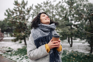 Young woman wearing frey fashion coat and blue scarf stands with a coffee to go at a winter snowy...