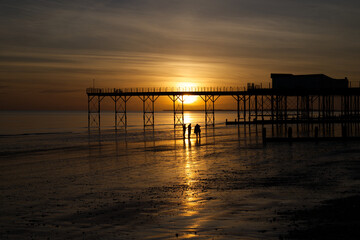 Fototapeta na wymiar A stunning sunset behind the Pier at Bognor Regis with beautiful reflections of the sinking sun in the sea and sand with locals admiring the spectacle. 