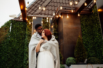 Obraz premium Portrait of beautiful couple wrapped in blanket stand at the balcony, handsome African American man hug gorgeous white woman, enjoy winter holidays, Christmas morning concept