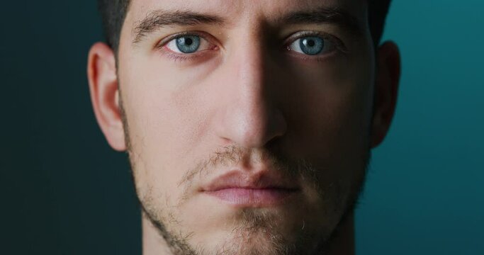 Cinematic close up of serious self confident young man with blue eyes is looking and blinking in camera isolated on dark background.