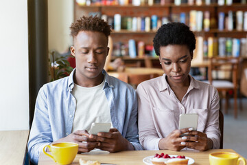 Gadget addiction and phubbing. Young black couple having dull date, stuck in smartphones, ignoring each other at cafe