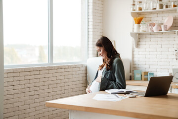 Joyful pregnant woman smiling while working with laptop and papers