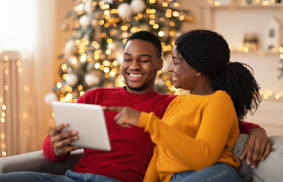Happy millennial lady points finger at tablet and shows to guy something interesting in interior with tree and garlands
