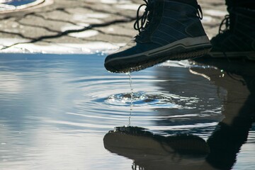 Foot in a shoe. Comes into a puddle. The blue sky is reflected in the water. White clouds. The sun shines