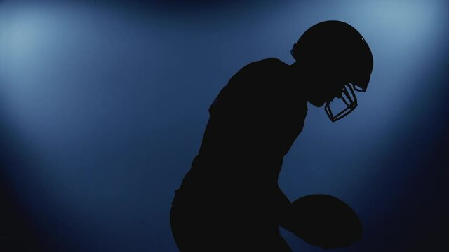 Silhouette of professional male American football player in helmet holding a ball walking against camera under bright stadium illumination lights. Usa team game and extreme sport spirit concept.