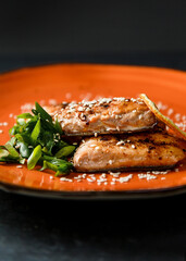 Grilled salmon served with green onions, orange and sesame seeds. Red fish on a large orange plate on a dark background, shot close up