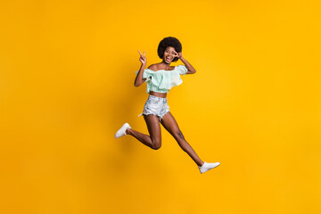 Fototapeta na wymiar Full length body size photo of jumping smiling black skinned girl showing v-sign fingers isolated on vivid yellow color background