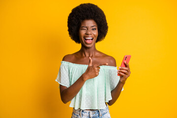 Photo portrait of winking girl holding phone in one hand pointing finger isolated on vivid yellow colored background