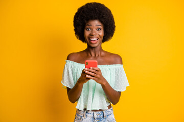 Photo portrait of excited girl with open mouth holding phone in two hands isolated on vivid yellow colored background