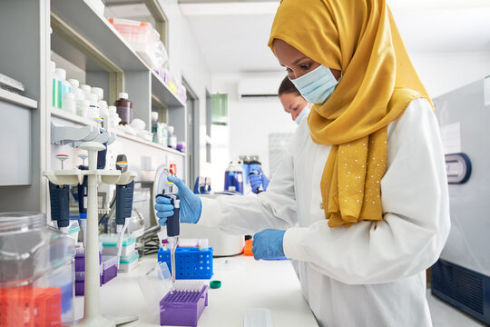 Female scientist in hijab and face mask using pipette in laboratory