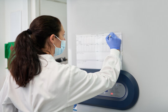 Female Scientist In Face Mask Marking Schedule On Wall In Laboratory