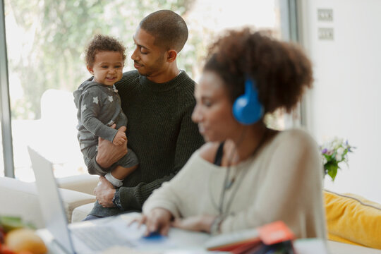 Father Holding Baby Daughter Behind Mother Working At Laptop