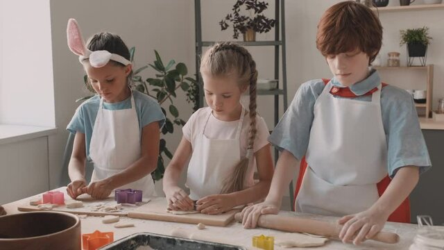 Waist up of red-haired boy rolling out dough with wooden rolling pin on table, Caucasian and Mixed-race school girls using cookie cutters. Children in aprons standing in kitchen, cooking