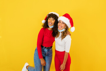 Beautiful latina curly brunette embracing her best female Caucasian friend smiling confident for future - Two mixed race women on yellow background in Santa hat wearing Coronavirus protection mask