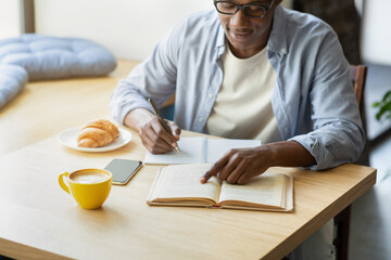 Cropped view of smart black guy taking notes from book at cafe, studying for test, copy space