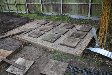 Close up of shed base construction of wood edging prepared hardcore compressed surface patio stone concrete slabs, a gravel surround french drain & brown earth soil & garden fencing background 