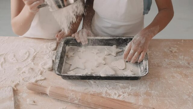 High angle shot of two unrecognizable people standing by kitchen table pouring flour through sifter over baking sheet with cookies in shapes of hearts and stars