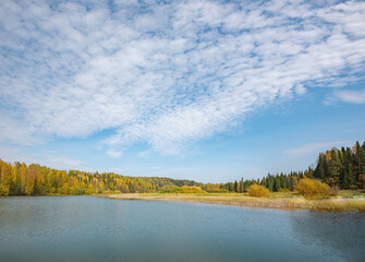 Autumn forest trees are reflected in the river water of the panoramic landscape. Blue sky with clouds.