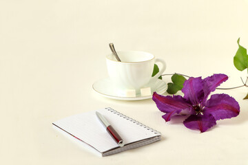 Notepad, pen, hot tea Cup with purple clematis flower on white background, side view, space for text