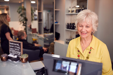 Senior Woman Arriving For Appointment To Have Hair Cut In Hairdressing Salon At Reception Desk