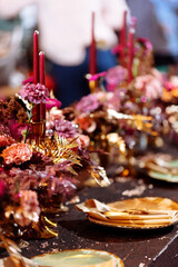 Christmas wedding dinner served table. Shiny leaves of gold paint, pink and red flowers and gold dishes. candles in a metal candlestick. Selected focus and blurred background