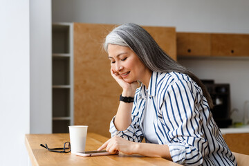 Happy white-haired mature woman drinking coffee and using mobile phone