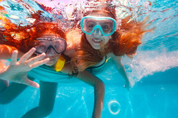 Two happy girls in scuba mask swim underwater and wave hand to the camera smiling in the pool