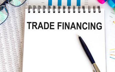 In the notebook, the text OF TRADE FINANCING, next to the pen, reports and glasses with graphs.