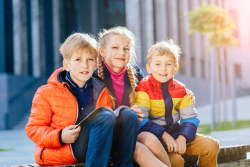 Group of three different age children sitting outdoor and reading book together. Two boys and girl after class in sunny autumn day. Urban background. Sun glare effect.