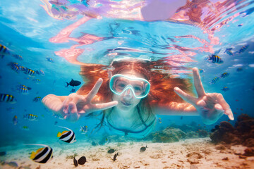 Portrait in scuba mask of the girl swim underwater among fish school and showing v victory gesture with hands