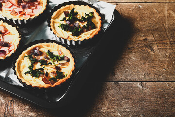 Mini individual quiche pies with filling ingredients ham,onion,spinich and cheese before egg is poured over the fillings in a baking tray on a rustic wooden background with a matt filter applied