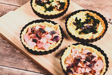 Mini individual quiche pies with filling ingredients ham,onion,spinich and cheese before egg is poured over the fillings in a baking tray on a rustic wooden background with a matt filter applied