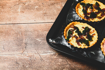 Mini individual quiche pies with filling ingredients ham,onion,spinich and cheese before egg is poured over the fillings in a baking tray on a rustic wooden background with a matt filter applied