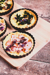 Mini individual quiche pies with filling ingredients ham,onion,spinich and cheese before egg is poured over the fillings in a baking tray on a rustic wooden background with a matt filter applied