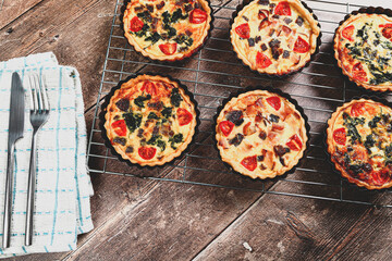 Freshly cooked mini individual quiche pies with filling ingredients ham,onion,spinich tomatoe and cheese cooling on a wire rack with knife and fork on a rustic wooden background with a matte filter 
