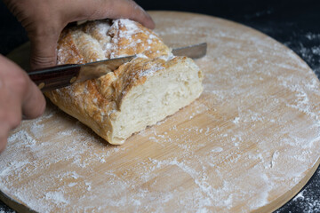 Person's hand cutting wheat traditional bread with a knife on floury wooden board.Rustic bakery concept.Dark background.