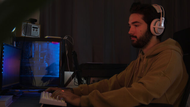Male using PC and concentrating while wearing white headphones in a darkened room