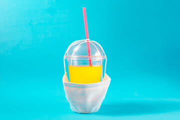 Yellow coctail in plastic cup with face mask on blue background. Quarantine catering concept.
