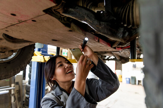 Smiling female auto mechanic using smart phone under car in garage
