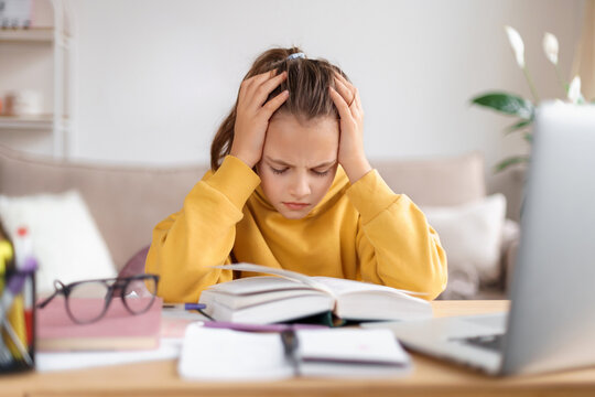 Exhausted schooler girl suffering from doing homework, tired from studying at home, getting ready before exams, stressed with difficult task, reading book, using laptop. Home education, homeschooling