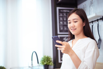 Cheerful Asian young woman reading an online news article and browsing the internet on a smartphone while relaxing in the kitchen. 