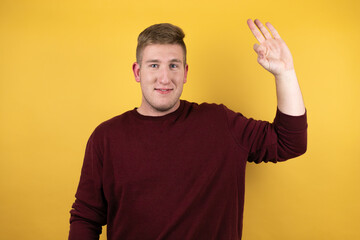 Young blonde man wearing a casual red sweater over yellow background doing ok sign with fingers and smiling, excellent symbol