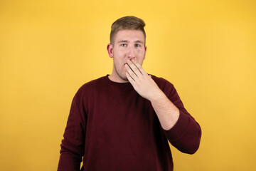 Young blonde man wearing a casual red sweater over yellow background surprised covering the mouth