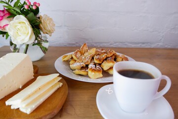Home-baked brushwood during quarantine. Homemade dessert. Homemade feta cheese, a cup of coffee on a wooden table top. Concept on the background of a white brick wall and a bouquet of roses.
