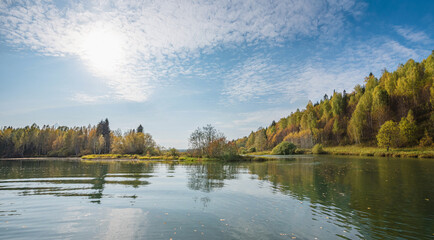 Autumn forest trees are reflected in the river water of the panoramic landscape. Blue sky with clouds.